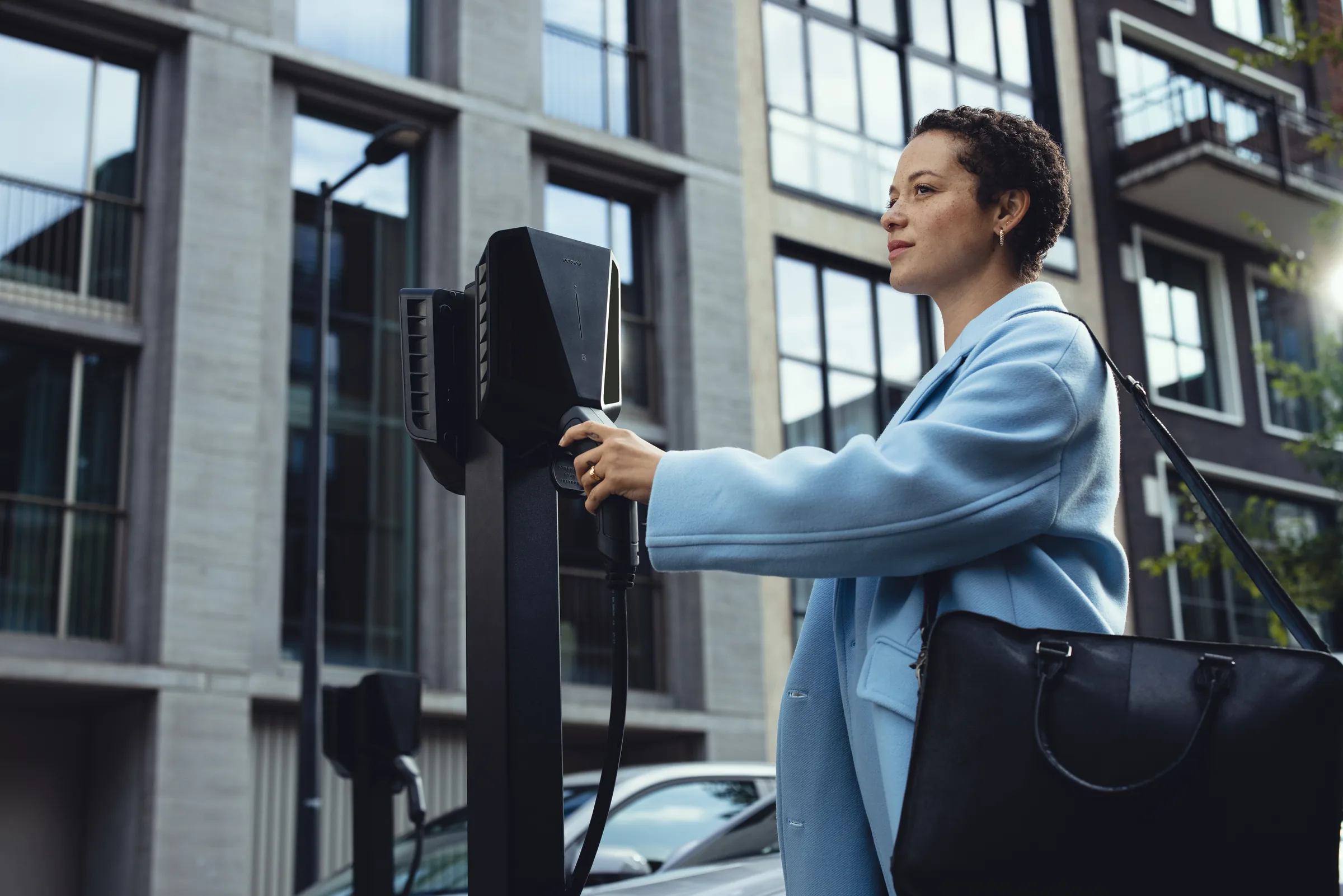 Woman in a light blue coat holding an RFID charging key up to a black Easee EV charger on a city street