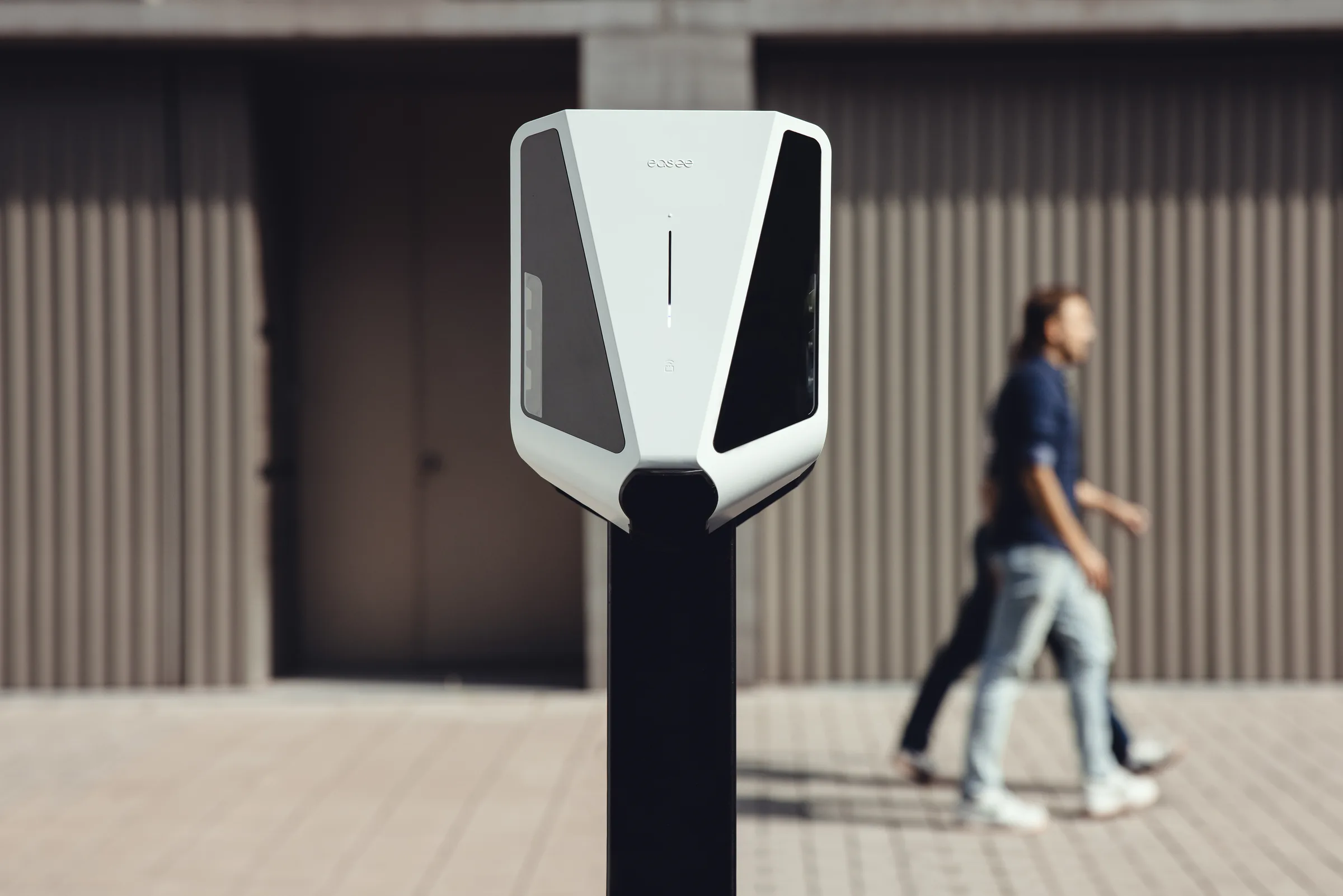 White Easee EV charger mounted on a black pole with pedestrians walking by in the background